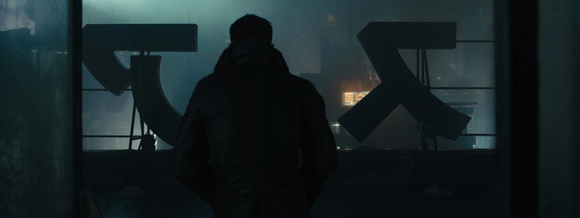 K at his apartment window, silhouetted against giant sign letters and a foggy city, from Blade Runner 2049.